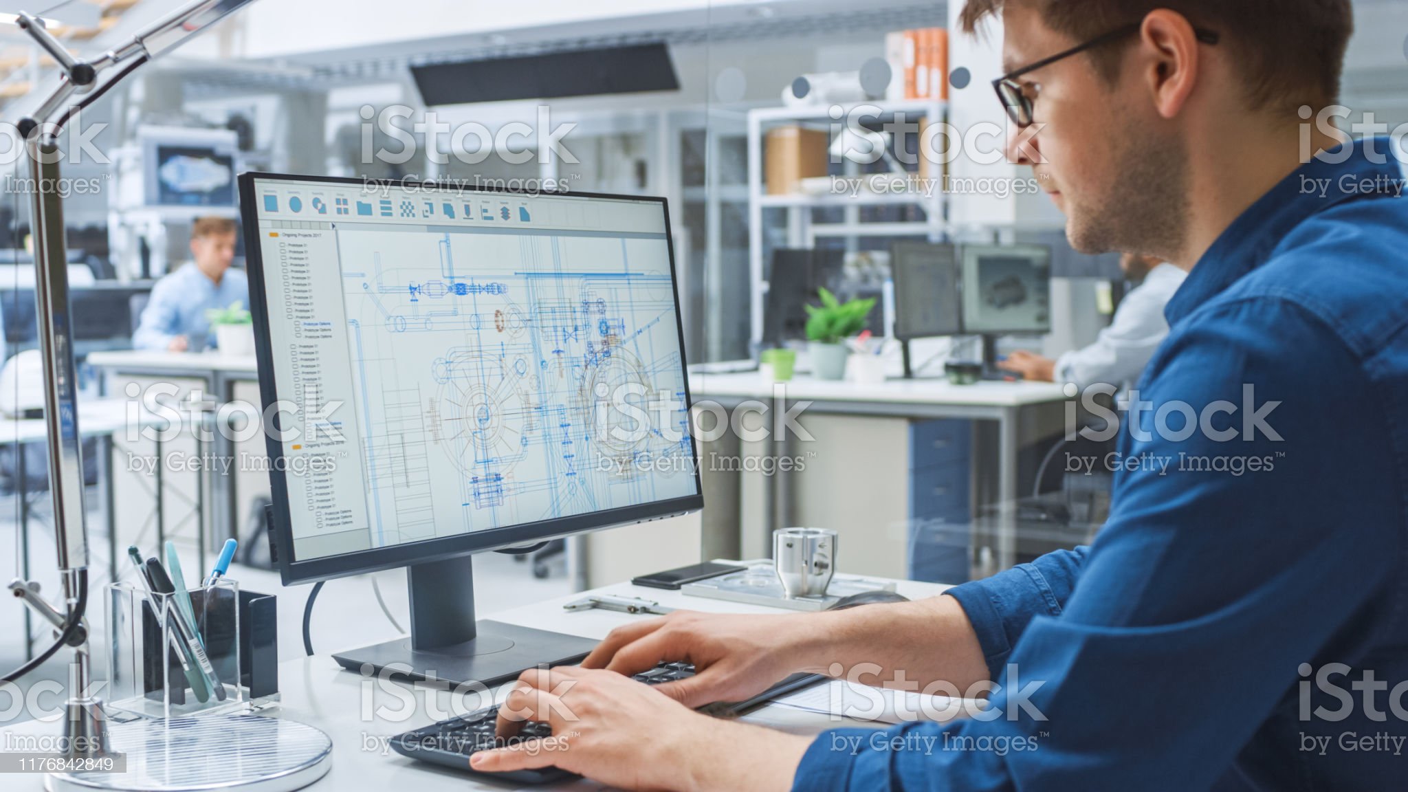 Over the Shoulder Shot of Engineer Working with CAD Software on Desktop Computer, Screen Shows Technical Drafts and Drawings. In the Background Engineering Facility Specialising on Industrial Design